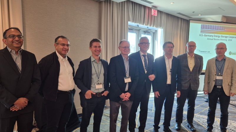 Acht Männer stehen in einer Reihe und lächeln in die Kamera, im Hintergrund ist eine Präsentationsfolie zur deutsch-amerikanischen Batterieforschungskooperation zu sehen. Die Aufnahme entstand beim Statusmeeting 2024 in Berkeley, Kalifornien