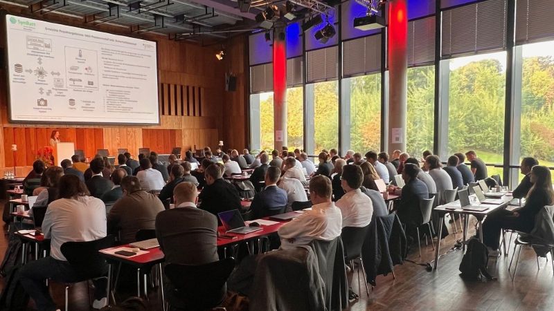Zahlreiche Menschen sitzen in Tischreihen in einem Veranstaltungsraumund schauen auf eine Präsentation auf einer Leinwand.