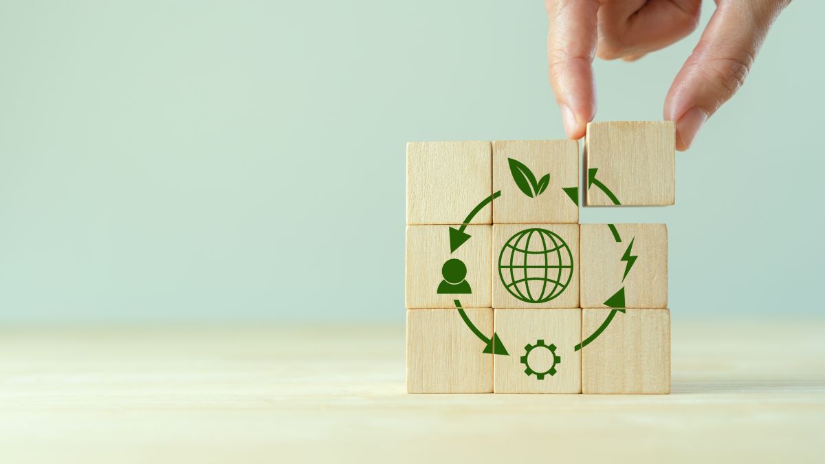 Eine Hand setzt den letzten Baustein in ein Puzzle aus Holzklötzen ein, das mit grünen Symbolen für Nachhaltigkeit, Energie und Umwelt gestaltet ist.